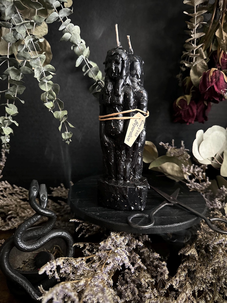 May include: A black sculpted candle with three figures, tied with a brown string and a tag that reads "Come Vibe Separation". The candle sits on a black wooden stand, with a metal snake and scissors nearby. Dried flowers and greenery surround the candle.