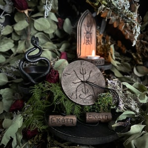 Może przedstawiać: Drewniany ołtarz z czarnym wężem, drewnianym świecznikiem ze świecą i drewnianym kołem z symbolem i napisem "Ekatm". Ołtarz otoczony jest suszoną zielenią.