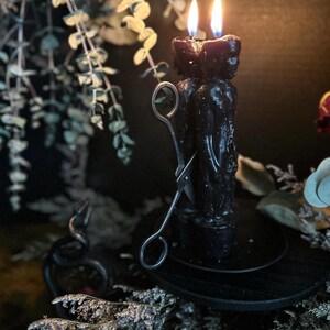 May include: A pair of black figure candles with lit flames, set on a black wooden stand. Silver candle snuffer rests nearby. The background features greenery and a decorative black snake, creating a mystical ambiance.