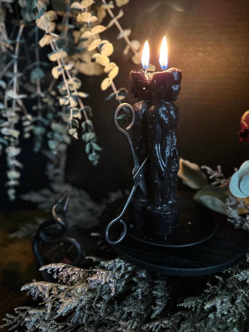 May include: A pair of black figure candles with lit flames, set on a black wooden stand. Silver candle snuffer rests nearby. The background features greenery and a decorative black snake, creating a mystical ambiance.