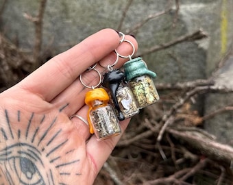 May include: Three miniature glass bottles filled with dried herbs, each with a different coloured stopper: orange, black, and green. The bottles are attached to silver keyrings. The background is a blurred outdoor scene with bare branches.