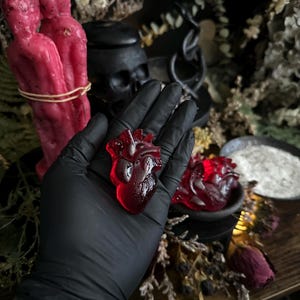 May include: A red, translucent anatomical heart-shaped object is held in a gloved hand. Two red figure candles are in the background, along with a black skull and other occult-themed items. The heart is approximately 2 inches in length.