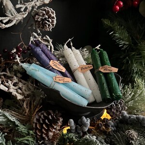 May include: A collection of spell candles in various colors, including purple, blue, white, and green, arranged in a dark metal bowl. Each candle has a small tag with text. The arrangement is surrounded by festive greenery and pinecones.