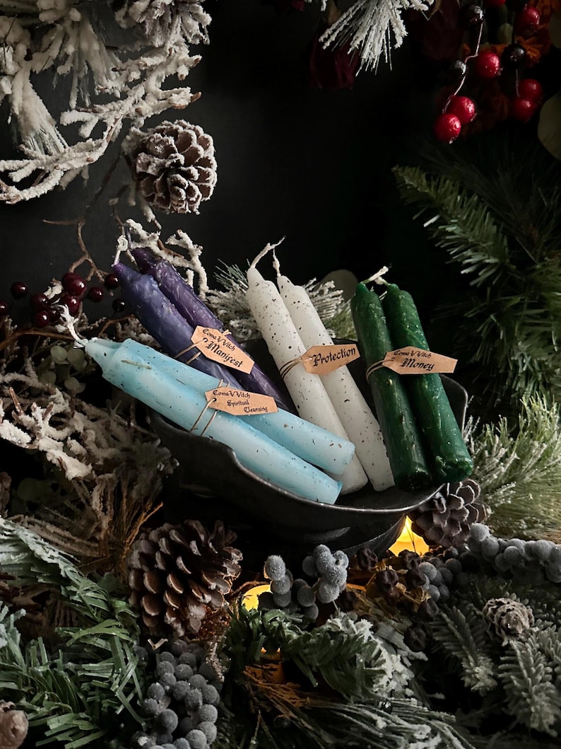 May include: A collection of spell candles in various colors, including purple, blue, white, and green, arranged in a dark metal bowl. Each candle has a small tag with text. The arrangement is surrounded by festive greenery and pinecones.