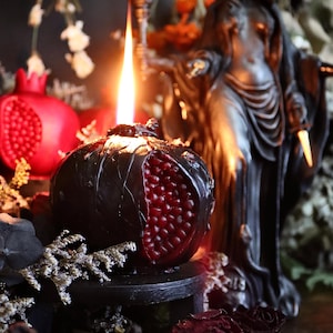 Puede incluir: Una vela encendida con forma de granada, con un exterior oscuro y un interior rojo, colocada sobre un pequeño soporte negro. Una granada roja y una estatua se encuentran en el fondo, junto con flores y follaje secos.