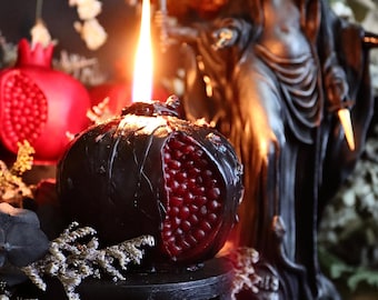 Puede incluir: Una vela encendida con forma de granada, con un exterior oscuro y un interior rojo, se encuentra sobre un soporte negro. Una estatua de una mujer con una diadema de media luna se alza en el fondo, junto a rosas secas y otros elementos decorativos.