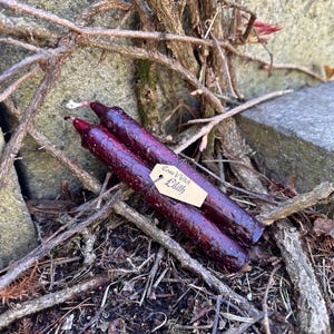 Puede incluir: Dos velas rituales de color burdeos oscuro con una superficie texturizada, atadas con hilo y una pequeña etiqueta que dice "Come Witch Lilith". Las velas están rodeadas de ramitas y ramas.