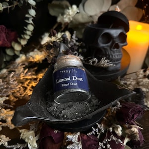 May include: A small glass jar labeled "Liminal Dust" sits in a black dish, surrounded by dried flowers and foliage. The jar has a cork stopper and a blue label. A black skull-shaped container is in the background, along with a lit candle.