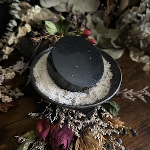 Puede incluir: Una piedra negra de forma ovalada descansa sobre un cuenco pequeño y oscuro lleno de gránulos blancos y hierbas. El cuenco se apoya en una base cuadrada oscura, rodeada de flores y follaje secos, creando una estética natural y terrosa.