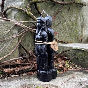 Può includere: Una candela nera raffigurante due figure, legate insieme con dello spago. La candela ha un'etichetta che recita "Come With Separation". La candela è posta su una superficie di pietra con muschio e viti sullo sfondo.