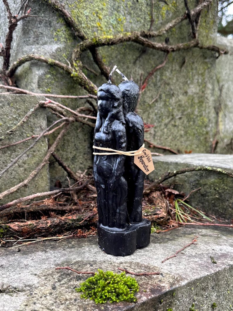 May include: A black candle depicting two figures, tied together with twine. The candle has a tag that reads "Come With Separation". The candle is placed on a stone surface with moss and vines in the background.