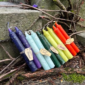 May include: A collection of seven colorful spell candles, each with a unique hue, including deep blue, purple, light blue, green, yellow, and red. Each candle is labeled with a small tag. The candles are arranged on a stone surface with natural foliage.