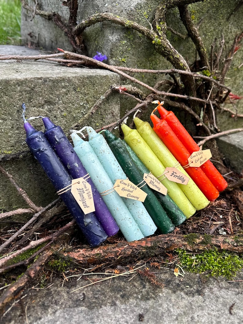 May include: A collection of seven colorful spell candles, each with a unique hue, including deep blue, purple, light blue, green, yellow, and red. Each candle is labeled with a small tag. The candles are arranged on a stone surface with natural foliage.