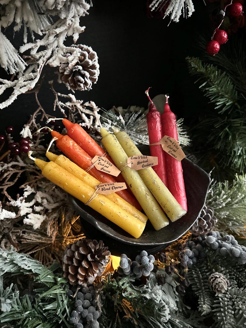 May include: A collection of colorful spell candles in a dark bowl, including yellow, orange, green, and red. Each candle has a small tag with text. The arrangement is surrounded by festive greenery and pinecones.