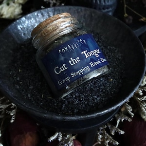 May include: A small glass jar with a cork lid, labeled "Cut the Tongue Gossip Stopping Ritual Dust." The jar sits in a dark, shallow bowl filled with black granules. The label is dark blue with white text.
