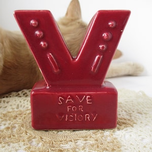 May include: A red ceramic letter "V" shaped figurine with the words "SAVE FOR VICTORY" inscribed on the base. The figurine has three raised dots on each side. The background includes a cat and a lace doily.