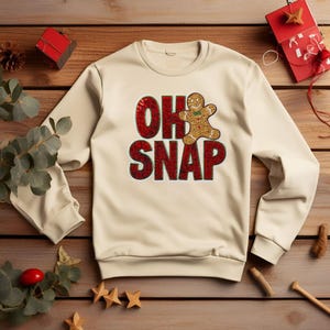 Può includere: Felpa color crema con la scritta "OH SNAP" in lettere rosse strutturate e una grafica di omino di pan di zenzero con un fiocco verde. La felpa è su una superficie di legno con decorazioni natalizie.