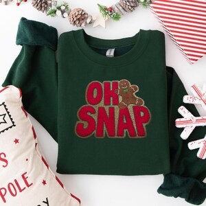 Könnte beinhalten: Ein dunkelgrünes Sweatshirt mit dem Schriftzug "OH SNAP" in Rot und einem Lebkuchenmann-Motiv. Das Sweatshirt hat einen Rundhalsausschnitt und lange Ärmel. Die Ärmel sind umgeschlagen, um ein dunkleres grünes Futter zu zeigen.