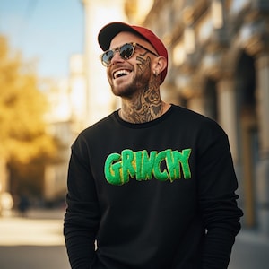 May include: A black sweatshirt with the word "GRINCH" in green and yellow lettering. The person in the image is wearing a red cap, sunglasses, and has tattoos on their face and neck.