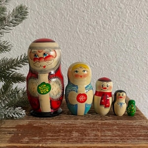 May include: A set of five Russian nesting dolls, also known as Matryoshka dolls, in a Christmas theme. The largest doll is Santa Claus, followed by a doll with a blue dress, a snowman, a penguin, and a small Christmas tree.