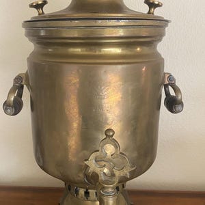 May include: Antique brass samovar with a cylindrical body, a spigot, and a lid with a small handle. The samovar has two handles on the sides and sits on a square base. The metal has a warm, aged patina.