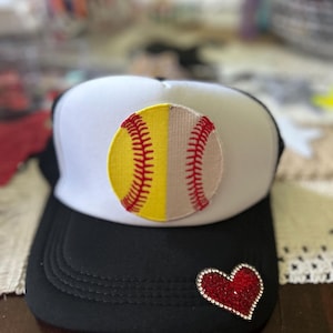 May include: Black and white trucker hat with a softball design. The hat features a split softball graphic in yellow and beige with red stitching. A rhinestone heart is on the brim. The hat has a white front panel and a black brim and mesh back.