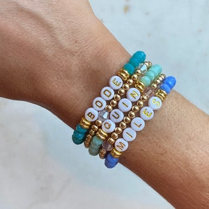 Puede incluir: Un conjunto de cinco pulseras de cuentas con cuentas blancas que deletrean las palabras "BOde", "QUIN", "MIL" y "NESS". Las pulseras están superpuestas en la muñeca de una persona y presentan cuentas de diferentes colores, incluyendo turquesa, azul claro y verde.