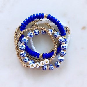 Puede incluir: Un conjunto de cinco pulseras con cuentas florales azules y blancas, cuentas doradas y una pulsera de silicona azul con la palabra "MAMA" escrita en letras doradas.