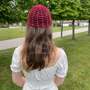 May include: A burgundy crochet hat with a grid pattern and small white beads. The hat is worn by a person in a white dress, standing in a park with green trees and grass.