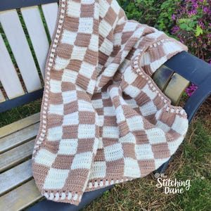 May include: A handmade crochet blanket with a checkerboard pattern in cream and brown. The blanket is draped over a park bench. The border has a decorative stitch. The blanket is made of soft yarn.
