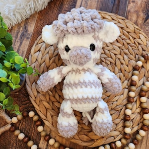 May include: A handmade crochet sheep toy in white and taupe. The sheep has a fluffy head, small ears, and black safety eyes. The body is striped with white and taupe yarn. The toy is sitting on a woven mat, with wooden beads and greenery in the background.