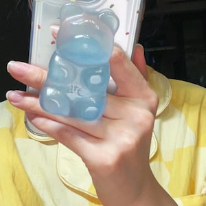 May include: A translucent blue gummy bear-shaped phone grip with the word "are" etched on its belly. The grip has a gradient effect, transitioning from a lighter blue at the top to a deeper blue at the bottom. The phone grip is held in a hand.