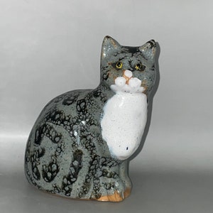 May include: A ceramic figurine of a grey and white cat with black spots. The cat is sitting with its head tilted slightly to the side and its eyes are closed.