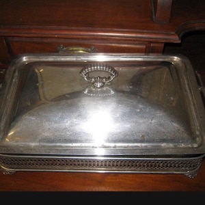 May include: A silver-plated chafing dish, rectangular with a domed lid and decorative handles. The dish has an openwork border and rests on small feet. The lid features a central handle. The design suggests a vintage or antique style, suitable for serving food.