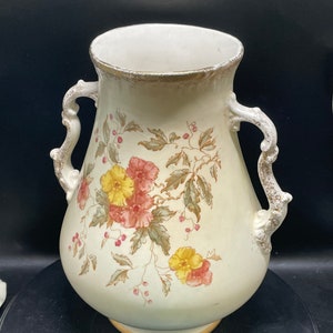 May include: A white ceramic vase with a floral design. The vase has two handles and a gold rim. The flowers are pink, yellow, and green.