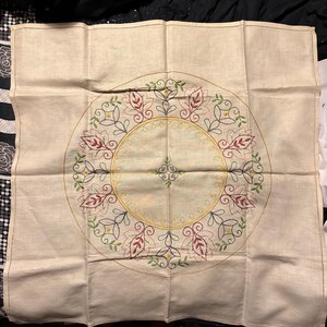 May include: A white linen tablecloth with an embroidered floral design in shades of red, green, yellow, and black. The design features a circle of leaves and flowers.