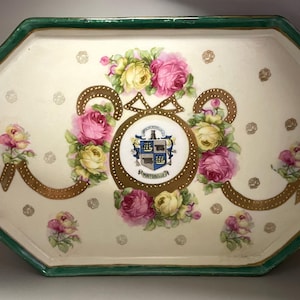 May include: An octagonal ceramic tray with a cream-colored base and a dark green rim. The center features a crest with the text "MONTICELLO" surrounded by a gold dotted ribbon and clusters of pink and yellow roses. Small gold floral accents.