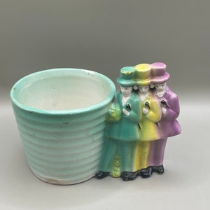 Puede incluir: Un macetero de cerámica verde, amarillo y morado con tres figuras que se toman de la mano. El macetero tiene un diseño a rayas y es perfecto para plantas pequeñas.
