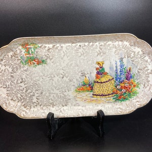 May include: A white oval serving tray with a gold rim and a floral pattern. The tray features a central image of a woman in a yellow dress standing in a garden with flowers.