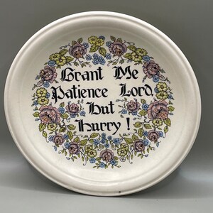 May include: A round, off-white ceramic plate featuring a decorative floral wreath of pink roses, yellow blossoms, and blue flowers. Centered within the wreath is the black text in a gothic-style font that reads: Grant Me Patience Lord, But Hurry!