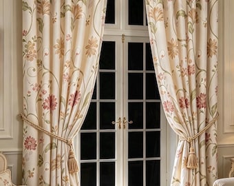 Puede incluir: Un par de cortinas de color crema que llegan hasta el suelo con un estampado floral en tonos rosa, naranja y verde. Las cortinas cuelgan de una barra dorada con pliegues y se sujetan con abrazaderas de cuerda trenzada con borlas.