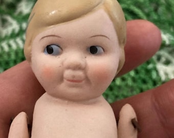Pode incluir: Uma boneca de porcelana com uma peruca castanho claro, olhos azuis e uma tez rosada. A boneca está nua e tem um leve sorriso.
