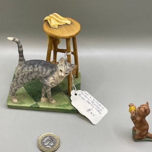 May include: Two ceramic cat figurines. One is a grey tabby cat standing on a green and white tiled base next to a wooden stool. The other is a brown tabby cat sitting on a green base and holding a yellow ball. The label reads "Cat & Kitchen Stool - Boulder Fine Arts 1984 #45".