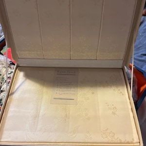 May include: A white box containing a cream-coloured tablecloth. The box has the words "Rayon Goods" printed on the front. The tablecloth has a floral pattern and the text "Instructions for Washing" is printed on a separate card inside the box.