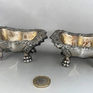 Op de afbeelding: Twee zilveren voetkommen met sierlijke details. De kommen zijn ovaalvormig en hebben een licht verweerde afwerking.