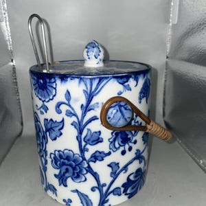 Puede incluir: Un cubo de hielo de cerámica blanca con un patrón floral azul y blanco y un asa de mimbre marrón. El cubo tiene una tapa con un pomo en la parte superior.