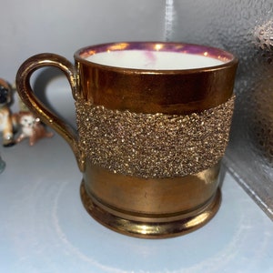 Könnte beinhalten: Eine Kupfertasse mit einem goldenen Glitzerband um die Mitte. Die Tasse hat ein weißes Innere und einen rosafarbenen Rand.