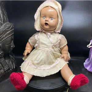 May include: A vintage porcelain doll wearing a white dress with pink trim and red knitted booties. The doll has a surprised expression with large blue eyes and a pink bonnet.