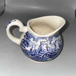 Puede incluir: Una jarra de crema de cerámica blanca con un diseño de castillo azul y blanco. La jarra tiene un asa y un cuerpo redondeado.