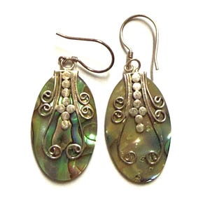 Op de afbeelding: Een paar ovaalvormige oorbellen met iriserende abalone schelp-inleg in groene en paarse tinten. Elke oorbel heeft een zilverkleurige metalen overlay met decoratieve krulmotieven en kleine ronde accenten, hangend aan een gebogen oorhaakje.